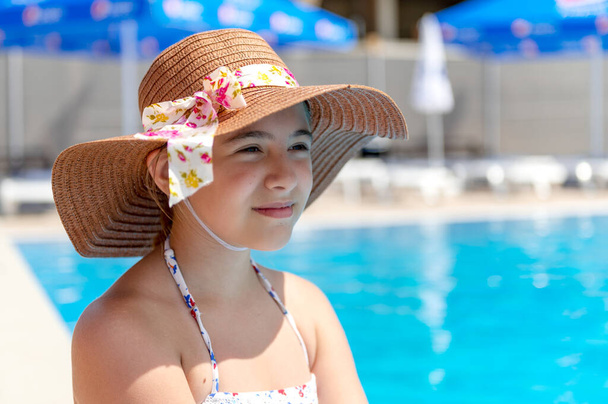 Hermoso sentado cerca de la piscina en un día soleado. Adolescente chica en un traje de baño y un sombrero se sienta cerca de la piscina del hotel con agua azul.  - Foto, Imagen