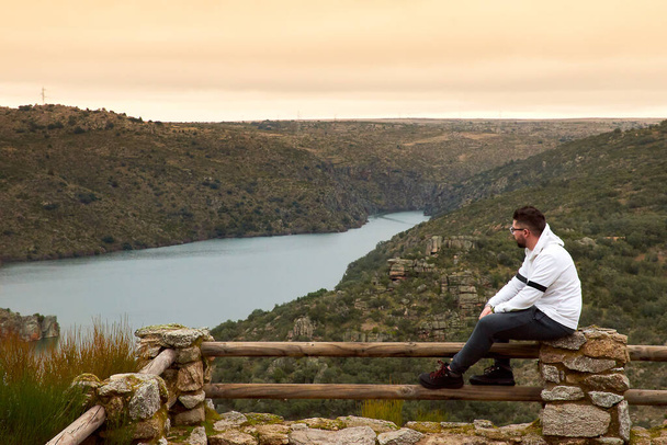 Homem branco caucasiano com óculos a olhar para o rio, sentado num mirante ao pôr-do-sol. Um montanhista de camisola branca. Trilho de madeira, lugar rústico. - Foto, Imagem