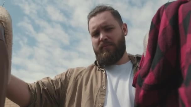  Alacsony szögű lövés szakállas férfi nézi használt zakók lóg ruházati állvány garázs eladó - Felvétel, videó
