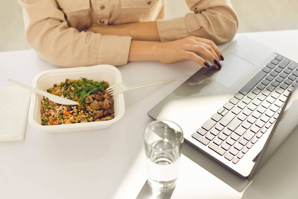 Businesswoman sitting at desk, having takeout lunch, drinking water and using laptop - 写真・画像