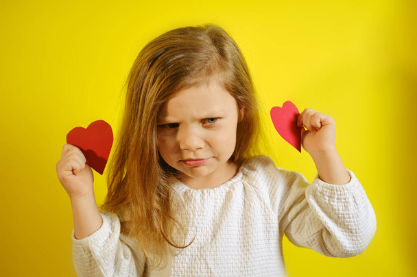 Portrait of upset or dissatisfied kid girl holding a red heart for you, isolated on yellow background with copy space. - Photo, image