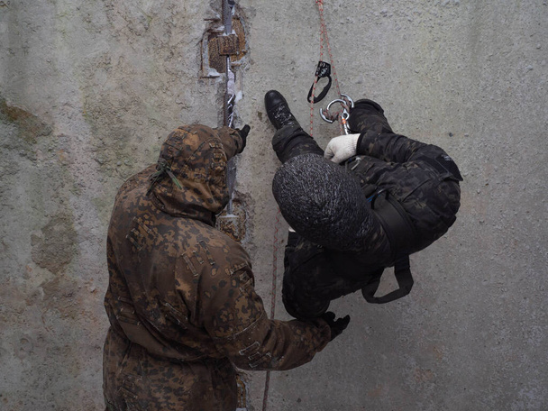 Ausbildung in Militär-, Sturm- und Polizeibergsteigen. Männlicher Instruktor bringt einem Mann in Tarnung bei, an einem Seil eine Wand hinunter zu klettern. Spezialeinheiten lernen auf dem Truppenübungsplatz. Foto ohne Gesicht. - Foto, Bild