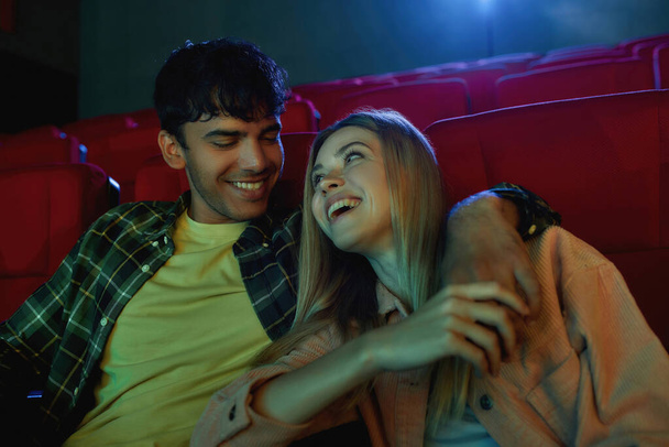 Πορτρέτο του ευτυχισμένου ζευγαριού, νεαρός άνδρας και γυναίκα κοιτάζοντας ο ένας τον άλλον, γελώντας μαζί, κρατώντας τα χέρια, ενώ βλέποντας ταινία, έχοντας ρομαντική ημερομηνία στο σινεμά - Φωτογραφία, εικόνα