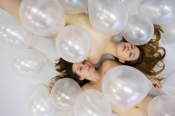 two attractive beautiful expressive young sexy smiling girls lying between white iridescent balloons on light grey ground floor - Photo, Image