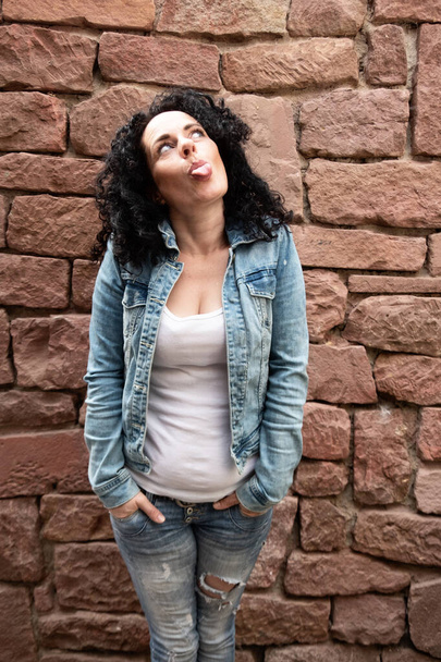 beauty sassy naughty mature woman with curly dark hair and white shirt and jeans jacket sticks out her tongue - Photo, Image