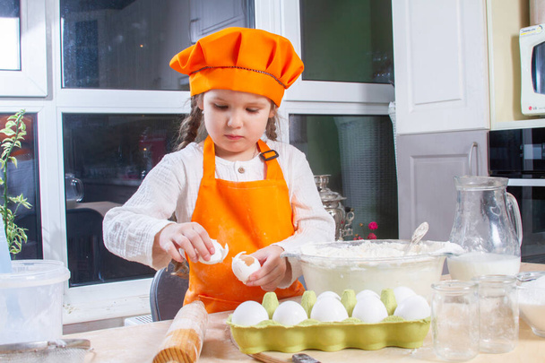 Маленькая симпатичная девочка в оранжевом костюме шеф-повара разбивает сырое яйцо и наливает его в миску муки, ребенок готовит и разминает тесто для выпечки, дочь помогает готовить на кухне. - Фото, изображение