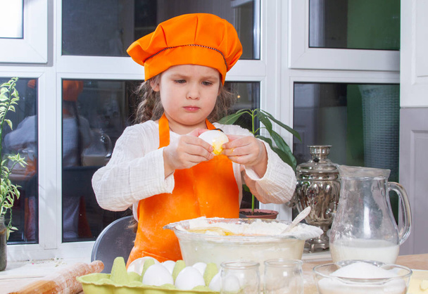 A little cute girl in an orange chef costume breaks a raw egg and pours it into a bowl of flour, a child prepares and kneads dough for baking, daughter helps to cook in the kitchen. - Fotoğraf, Görsel