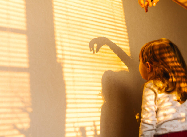 Selective focus. Time spent during quarantine. Sun and shade. Little girl in pajamas at home playing with the sun on the wall. Photo from behind. - Foto, immagini