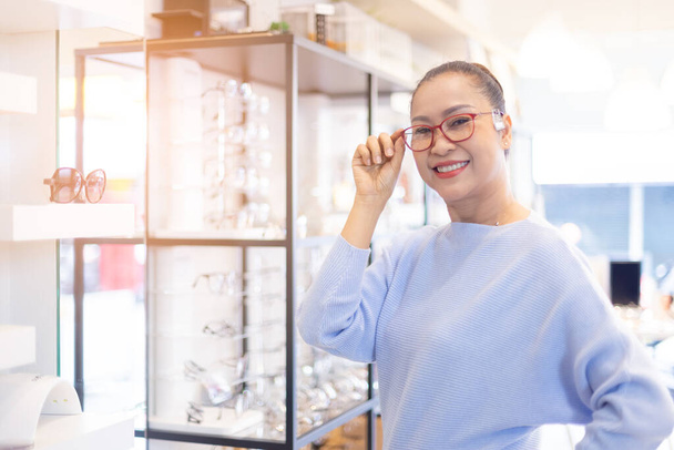 Donna asiatica di mezza età che sceglie occhiali occhiali in negozio ottico. usato corretto o assistere la vista difettosa, sorridente e guardando la fotocamera. - Foto, immagini