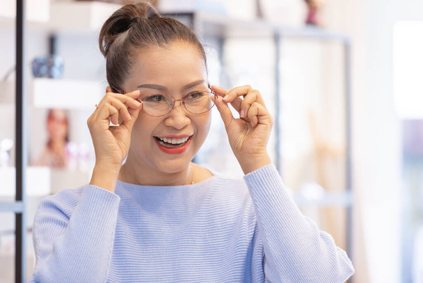 Asiatin mittleren Alters wählt Brille im Optikergeschäft. Korrektes oder fehlerhaftes Sehvermögen unterstützen. - Foto, Bild