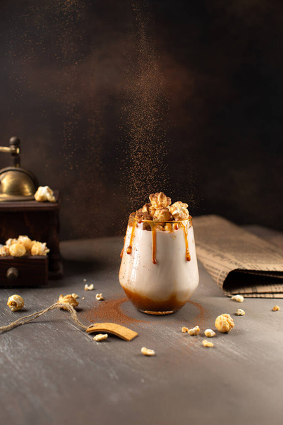 Sweet Milkshake with caramel syrup,cream liqueur,caramel popcorn and chocolate powder on brown background with vintage,manual coffee grinder and falling cocoa powder. - Fotoğraf, Görsel