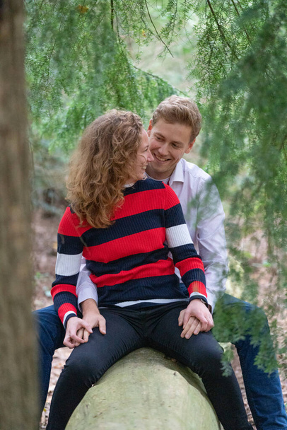 Beautiful enamored young couple sitting on a tree trunk in the forest. Facial expression is in love. In half body, man sits behind the woman - Photo, Image