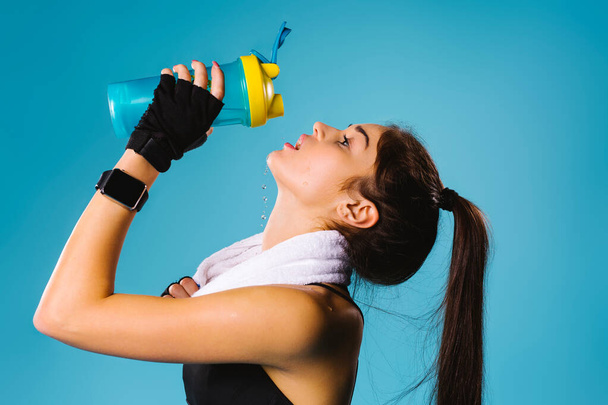 Sporty brunette girl after a workout with a towel on her neck drinks protein shakes. Blue background. - Foto, imagen