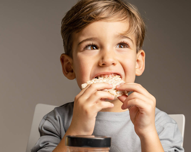 小さな白人の少年の肖像画を閉じます4歳食べるクリスピーパフライスケーキ横に見える-健康的なグルテンフリービーガンやベジタリアン料理-フロントビュースタジオショット - 写真・画像