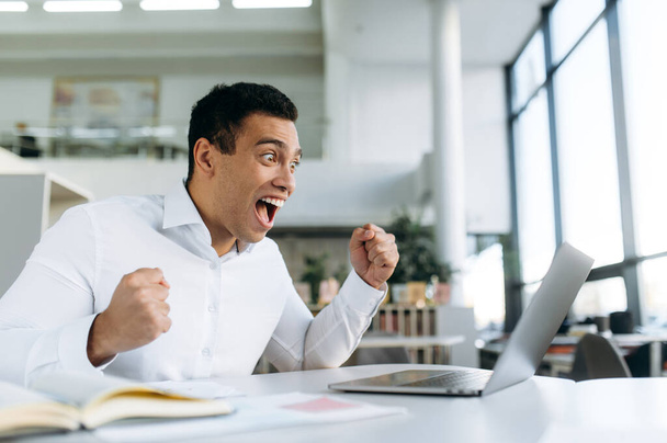 Surprised hispanic male sitting at the work desk, with excited face, receive pleasant news. Overjoyed excited man, got a good job promotion or good news, success concept - Photo, Image
