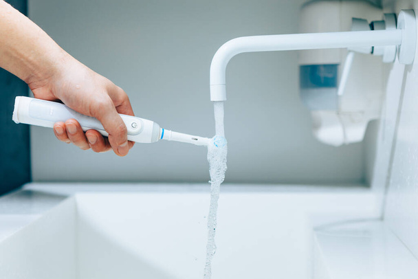 hand houden van een tandenborstel onder stromend water van kraan in een badkamer - Foto, afbeelding