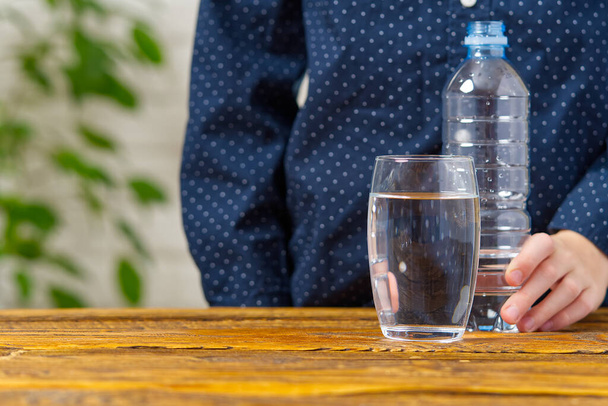 Мальчик наливает воду из пластиковой бутылки в стекло на деревянный стол - Фото, изображение