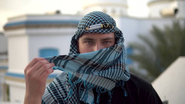 Jongeman in Arabische zakdoek. De man bedekt zijn gezicht. Tegen de achtergrond van het Arabische huis. - Foto, afbeelding