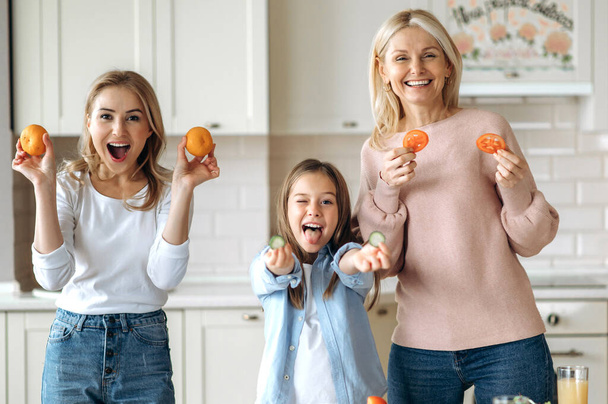 Three female generation spend time together at the kitchen at home. Caucasian happy grandma daughter and granddaughter cook together a fresh salad, indulge with vegetables, fruits and smiling - Photo, Image