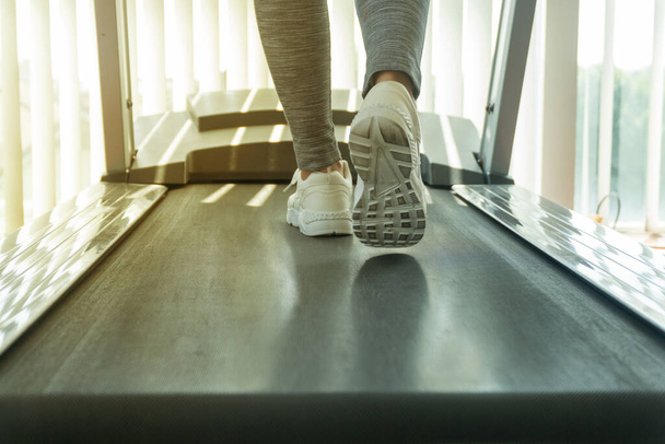 Junge Frau mit muskulösen Füßen in Turnschuhen, die auf dem Laufband im Fitnessstudio mit Sonnenlicht-Effekt laufen. Trainings- und Sportkonzept - Foto, Bild