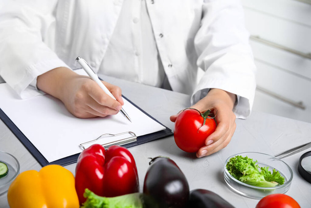 Scientist with clipboard and fresh tomato at table in laboratory, closeup. Poison detection - Fotó, kép