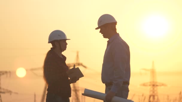 Engineers man and woman work on the field with electric towers at sunset. Teamwork of engineers, discussion and coordination of work. High voltage power plant business project. Handshake, agreed. - Footage, Video