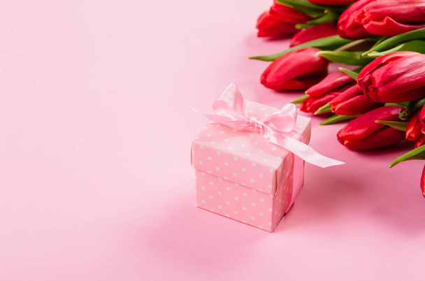 Gift box with a pink ribbon in white polka dots on a background of a bouquet of red tulips with copy space for text. Wallpaper or banner for gift shop, flower shop or jewelry store. Valentine day. - Fotó, kép