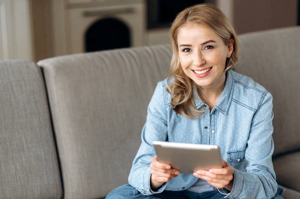 Kaukasische attraktive blonde Frau in Freizeitkleidung sitzt auf der Couch, nutzt Tablet, surft im Internet, sozialen Netzwerken, chattet mit Freunden, shoppt online - Foto, Bild