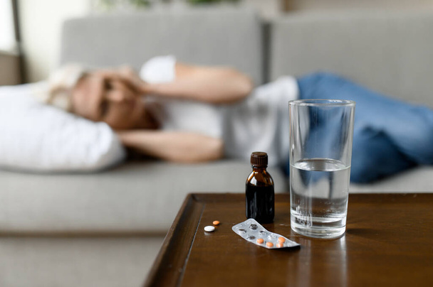 Caucasian unhealthy mature defocused woman lying on sofa with headache due to illness, virus, high temperature, need a rest sleep and pills. A glass of water and medicine on the table - Foto, immagini