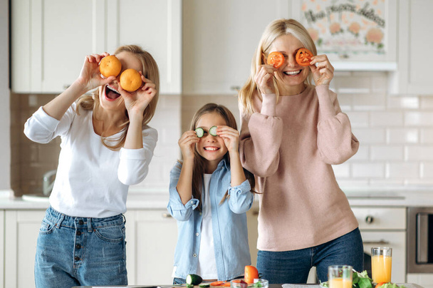 Drie vrouwelijke generaties brengen samen tijd door in de keuken thuis. Kaukasisch gelukkig oma dochter en kleindochter koken samen een verse salade, genieten van groenten, fruit en glimlachen - Foto, afbeelding