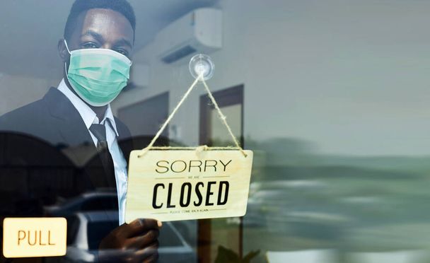 Ένας μαύρος σερβιτόρος που φοράει μάσκα κατά του ιού κρατά μια πινακίδα που λέει ότι το μαγαζί είναι κλειστό λόγω της μεγάλης επιδημίας του κορωναϊού..  - Φωτογραφία, εικόνα