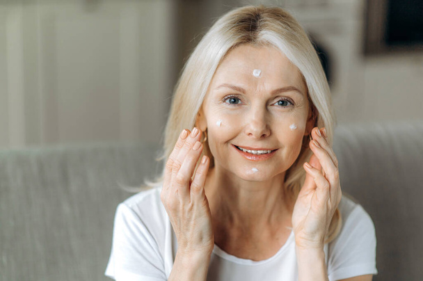 Oudere huidverzorging. Aantrekkelijke blanke dame van middelbare leeftijd zorgt voor haar gezicht, brengt anti-aging rimpel crème, kijkt naar de camera en glimlacht. Close-up - Foto, afbeelding