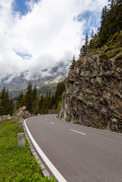  spring view of Swiss Alps at high elevation. Clouds move by the summits of steep cliffs and mountains. Pine trees and grass are seen alongside a curvy mountain road. - Photo, Image