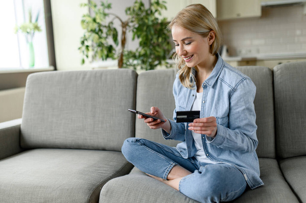 Online shopping, online payment. Joyful caucasian blonde woman dressed in casual clothes, shopping online while sitting in living room using credit card and smartphone - Foto, Imagen