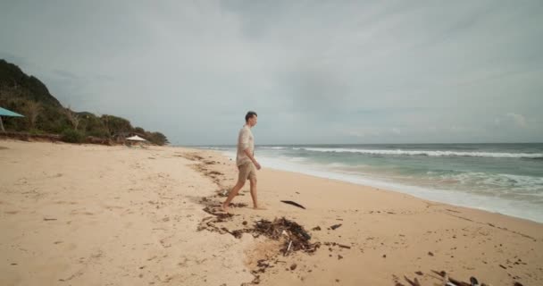 Joven hombre blanco en pantalones cortos de color caqui y camisa caminando hacia el océano en la hermosa playa tropical de Bali con algas marinas - Metraje, vídeo