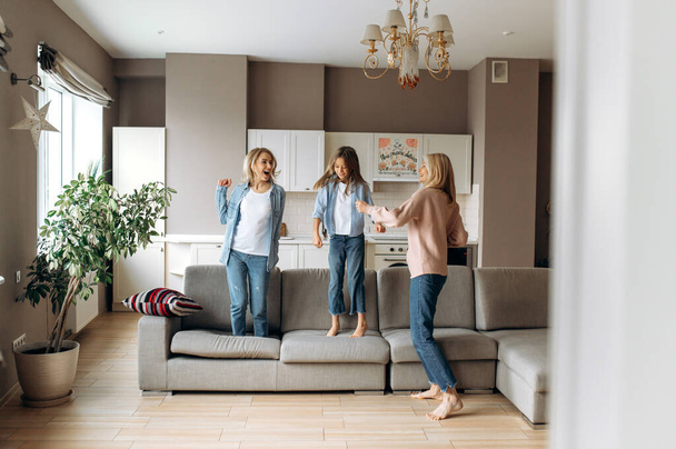 Cheerful caucasian mother, daughter and modern grandmother are having fun at home together, they are dancing, jumping on the sofa, fooling around and laughing - Foto, Bild