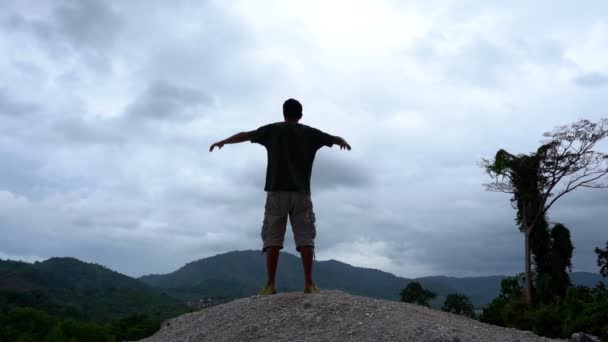 Nuori aasialainen mies kurottautuu vuoristoalueen huipulle, seisoo vuoren huipulla ja nostaa kätensä ylös. tietoisuus menestyksestä hidastettuna - Materiaali, video