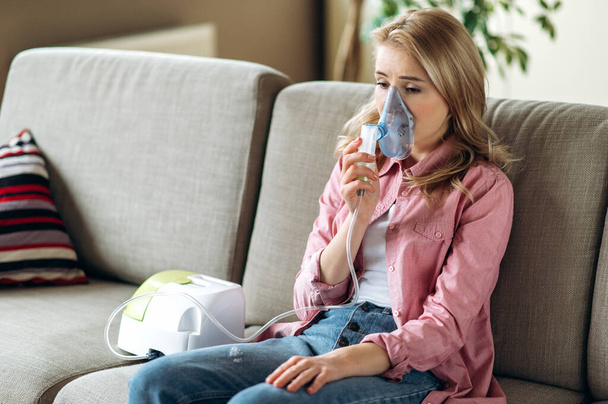 Una mujer caucásica preocupada con un inhalador. Hembra poco saludable haciendo inhalación en casa, usa nebulizador e inhalador para el tratamiento sentado en el sofá en casa - Foto, imagen