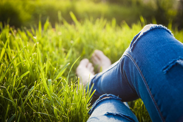 引き裂かれたジーンズを着たヒップスター女性の足が緑の草の中に横たわっていますスマートフォンとヘッドセット日没を楽しんで-夏のリラクゼーション - 写真・画像