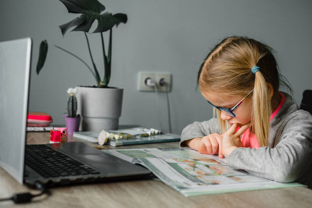 Menina em óculos olhando para o seu livro de aula, tendo aula on-line em casa - Foto, Imagem