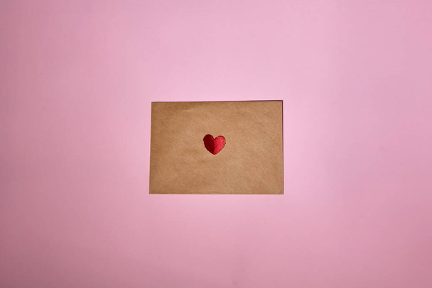 A red heart lies on a brown envelope for Valentine's Day. Pink backdrop with envelope and red heart. Love letter or message concept. Flat-lay, top view. Copy space for your text.  - Foto, Imagen