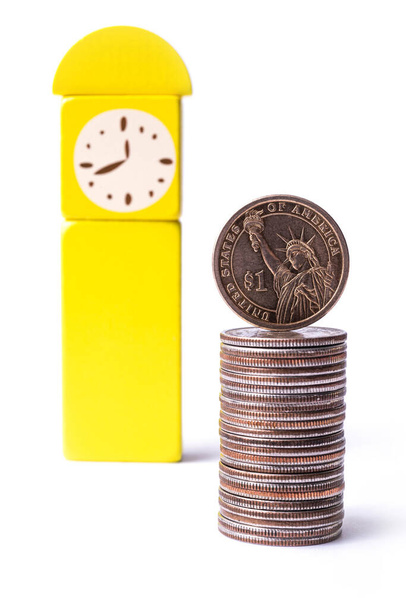 Een munt in denominatie van een Amerikaanse dollar staat op de rand van een stapel van 25 cent naast een klok gemaakt van kinderen gele kubussen op een witte achtergrond - Foto, afbeelding