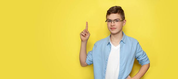 The boy is a teenager wearing glasses, a blue shirt and a white T-shirt on a yellow background. The boy calls for attention, raising his finger up. A place for text. - Foto, Imagem
