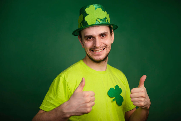 Um homem com um chapéu de trevo e uma t-shirt verde mostra o polegar para cima. Classe de gestos. O tipo está a celebrar o dia de São Patrício. Estúdio foto - Foto, Imagem
