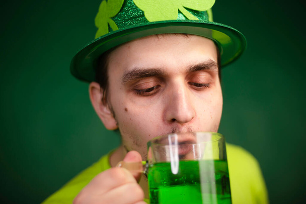 A man in a shamrock hat and green T-shirt drinks a large mug of green ale. Guy celebrates st patrick's day with a glass of beer. - Фото, зображення