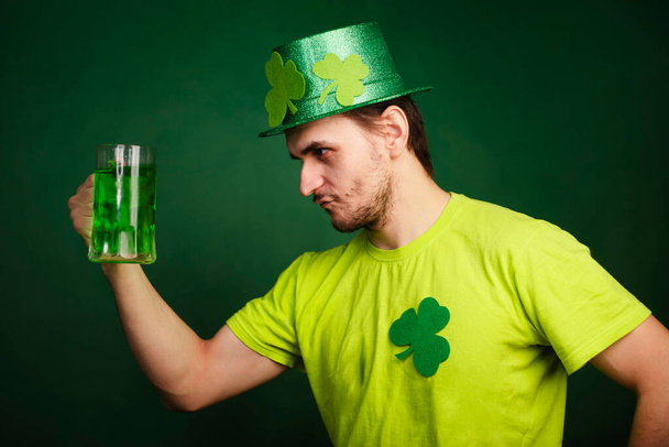 Мужчина в шляпе и зеленой футболке пьет большую кружку зеленого эля. Парень празднует День Святого Патрика с бокалом пива. - Фото, изображение