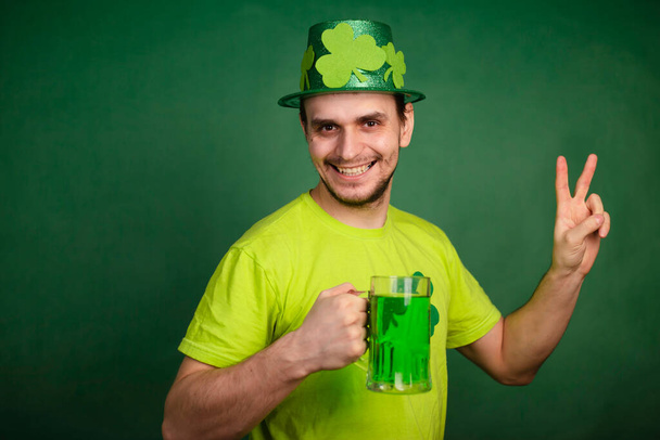 Um homem de chapéu de trevo e t-shirt verde bebe uma grande caneca de cerveja verde. Guy comemora o dia de São Patrício com um copo de cerveja. - Foto, Imagem