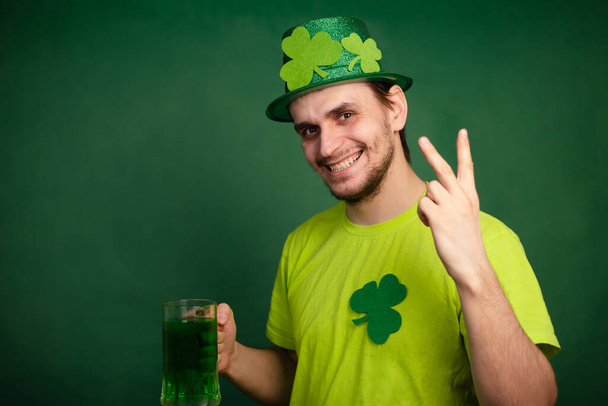Un hombre con un sombrero de trébol y una camiseta verde bebe una taza grande de cerveza verde. Guy celebra el día de San Patricio con un vaso de cerveza. - Foto, imagen