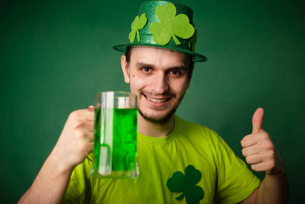 Ένας άντρας με καπέλο τριφύλλι και πράσινο μπλουζάκι πίνει μια μεγάλη κούπα πράσινη μπύρα. Ο τύπος γιορτάζει τη μέρα του Αγίου Πατρικίου με ένα ποτήρι μπύρα.. - Φωτογραφία, εικόνα