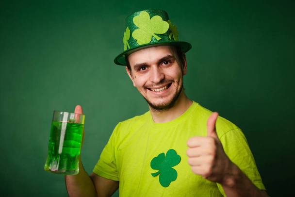 Un hombre con un sombrero de trébol y una camiseta verde bebe una taza grande de cerveza verde. Guy celebra el día de San Patricio con un vaso de cerveza. - Foto, imagen